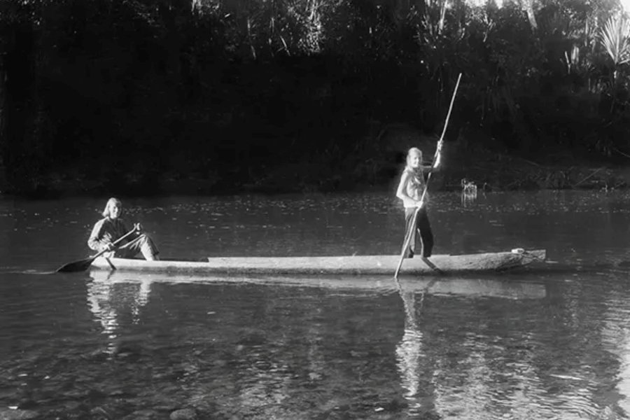 Two people in a dugout canoe navigate a calm river; one sits paddling while the other stands with a long pole, possibly steering. The scene is in black and white, with dense forest in the background and the water reflecting their movements.