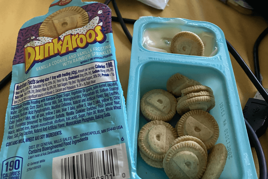 A pack of Dunkaroos featuring vanilla cookies in a tray alongside a separate compartment of vanilla frosting with rainbow sprinkles. The packaging is visible and labeled with nutrition facts, ingredients, and branding in colorful retro-style font.
