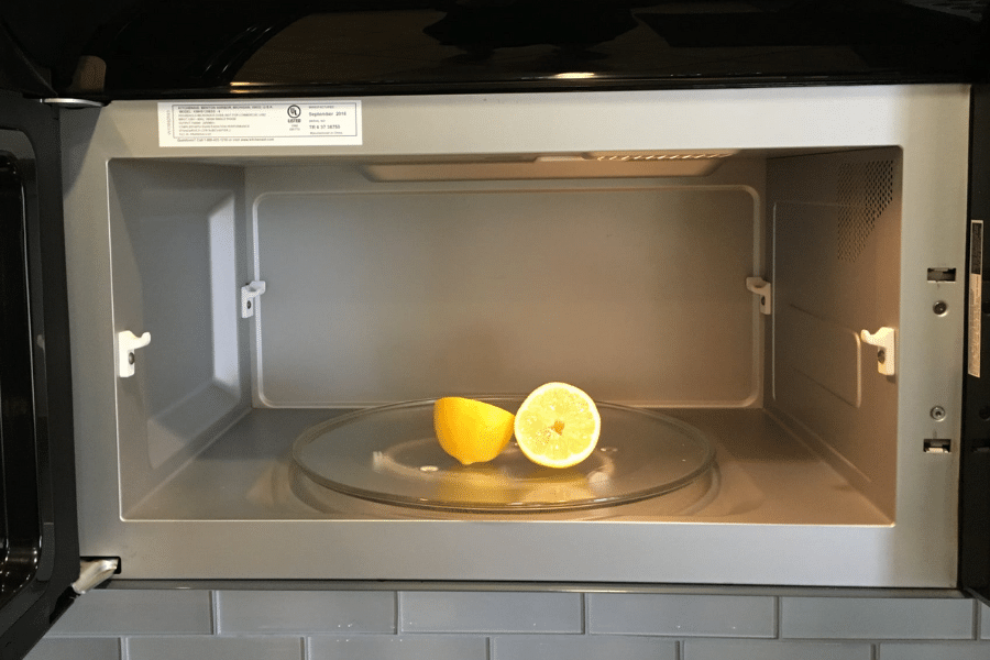 Open microwave with a halved lemon placed on the glass turntable, positioned for steam cleaning or deodorizing. The microwave interior is clean, with a visible label on the top edge and a tiled backsplash underneath.