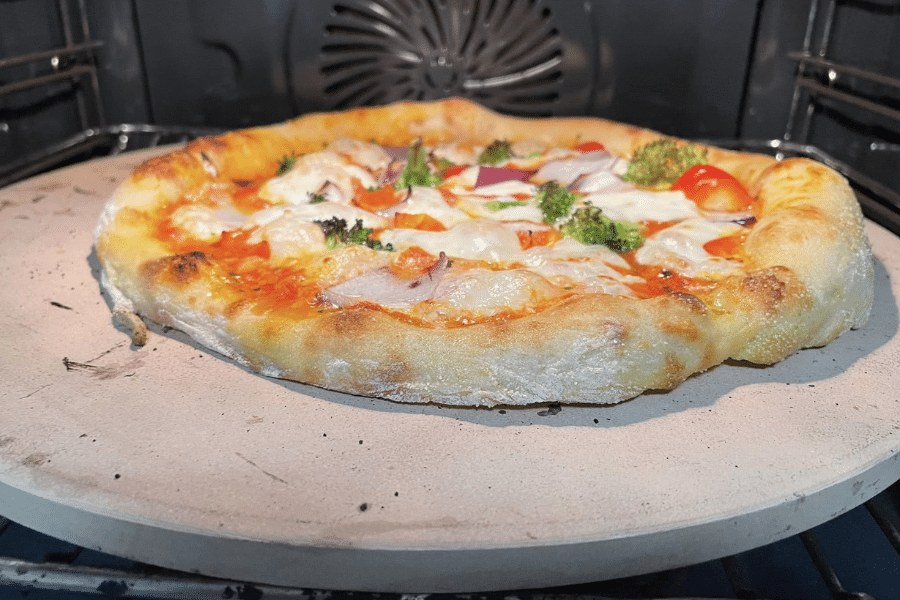 Freshly baked homemade pizza with a golden, puffy crust and toppings like mozzarella, broccoli, red onion, and cherry tomatoes, sitting on a pizza stone inside an oven.