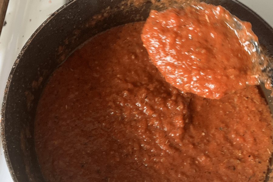 Close-up of a pot filled with thick, chunky red pasta sauce, with a spoon scooping some of it up. The sauce appears rich and seasoned, likely made from tomatoes, herbs, and spices.