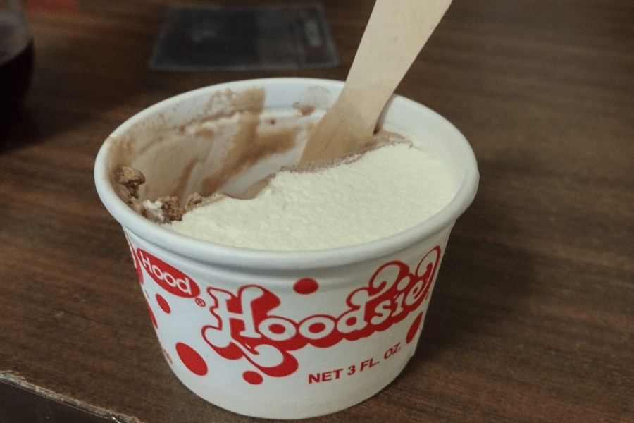 A small 3 oz paper cup of Hood Hoodsie ice cream sits on a wooden surface with a wooden spoon sticking out. The ice cream is a classic vanilla and chocolate swirl, with a portion already eaten, revealing the smooth texture inside.