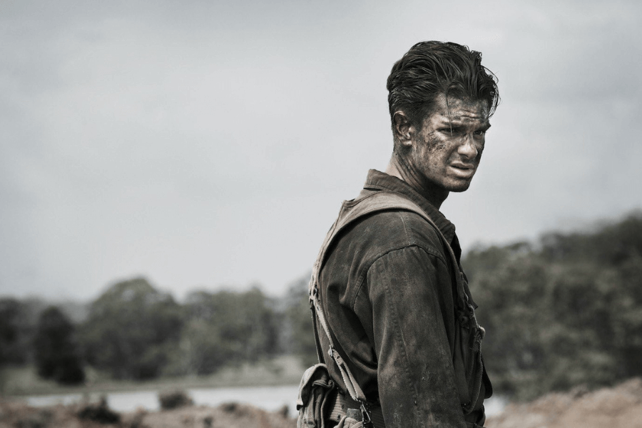 Scene from a war film showing a soldier covered in dirt and grime, wearing a worn military uniform and gear, standing against a desolate, cloudy backdrop. His intense expression and battle-worn appearance reflect the emotional toll of combat.