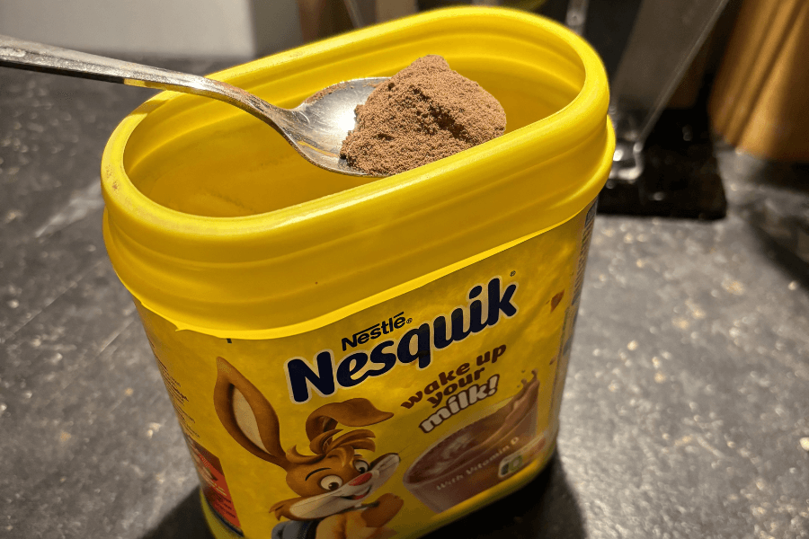 A yellow container of Nestlé Nesquik chocolate powder sits open on a kitchen counter, with a spoonful of the cocoa powder held above it. The label features the Nesquik bunny and the slogan "wake up your milk!"