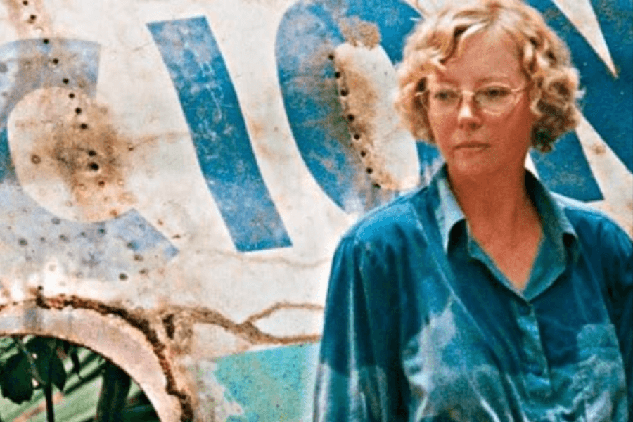 Close-up of a woman with curly blonde hair and glasses standing in front of a rusted, faded aircraft fuselage with large blue letters partially visible. She wears a worn teal shirt, and her expression is somber and introspective.