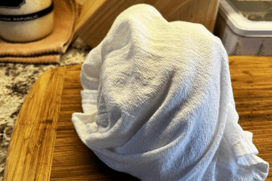 A white kitchen towel draped over a rounded object—likely a bowl of dough—on a wooden cutting board, suggesting a dough resting or proofing process. The scene includes kitchen tools and containers in the background.