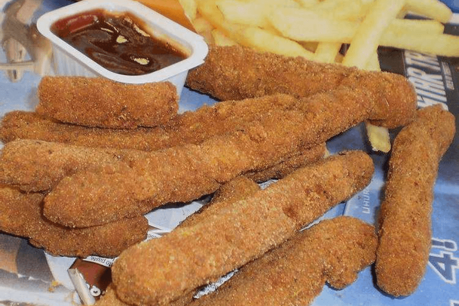 A fast food meal featuring a pile of crispy, breaded chicken fries served on a tray liner, with a small container of dark dipping sauce and a portion of French fries in the background.