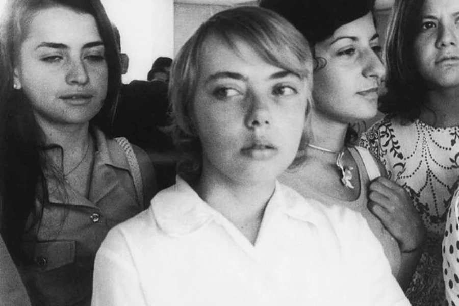 Black and white photo of a young woman in a white blouse looking off to the side, surrounded closely by other young women in a crowded indoor setting. Their expressions range from calm to contemplative, suggesting a serious or intense moment.