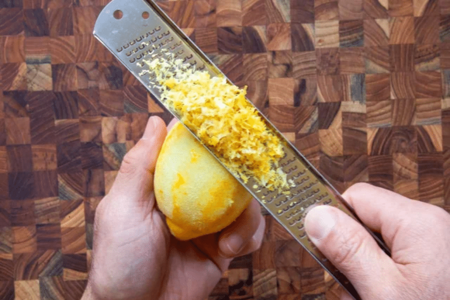 Close-up of hands zesting a lemon using a fine grater over a wooden butcher block surface. A pile of fresh yellow lemon zest collects on the grater, highlighting the citrus aroma being prepared for use.