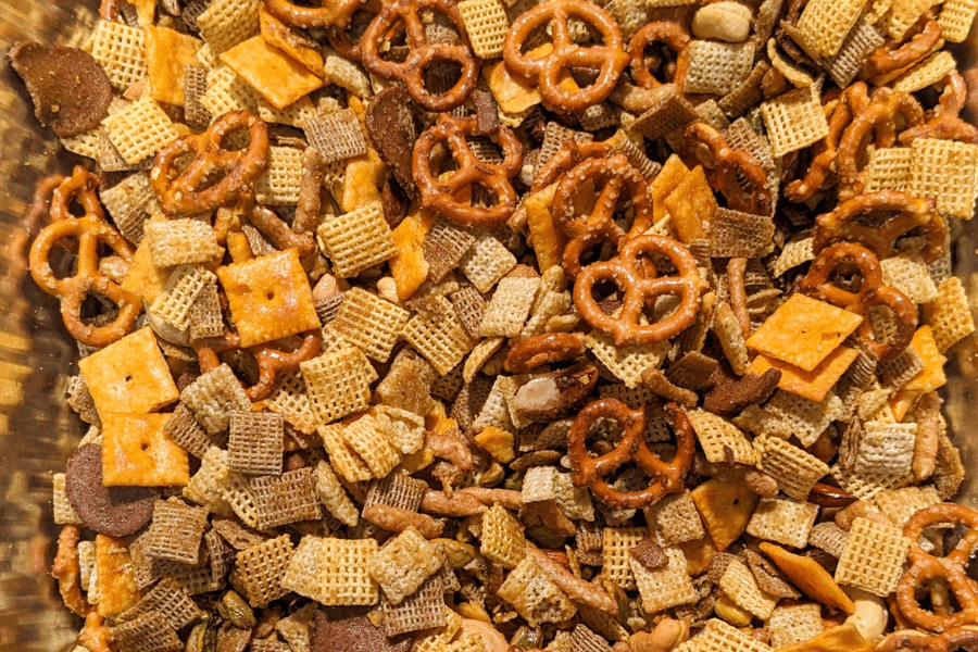 A close-up of a homemade snack mix featuring a variety of ingredients including mini pretzels, Chex cereal, cheese crackers, rye chips, and assorted nuts. The mix is golden and crunchy with a savory seasoning coating.