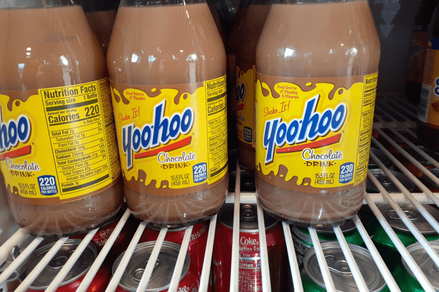 Glass bottles of Yoo-hoo chocolate drink are lined up in a refrigerator, with the yellow and brown label reading “Shake It!” and “Good Source of Vitamins & Minerals.” The bottles sit above cans of soda visible through the fridge shelf bars.
