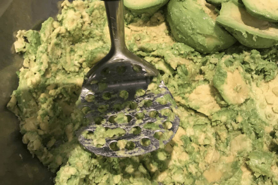 Close-up of a metal potato masher pressing into a bowl of ripe avocados, in the process of being mashed for guacamole or a similar dish. The texture is chunky with some whole avocado halves visible.