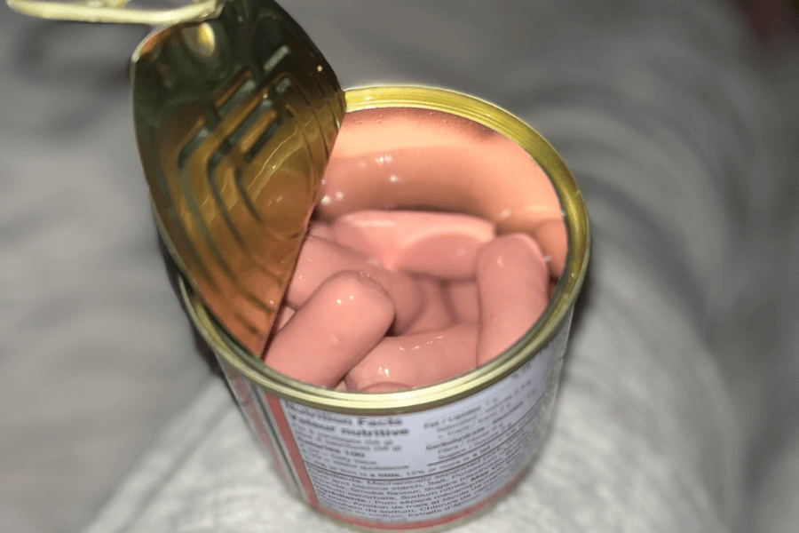 An opened can filled with pale pink cocktail sausages submerged in a clear brine or liquid. The metal lid is partially peeled back, revealing the sausages packed tightly inside.