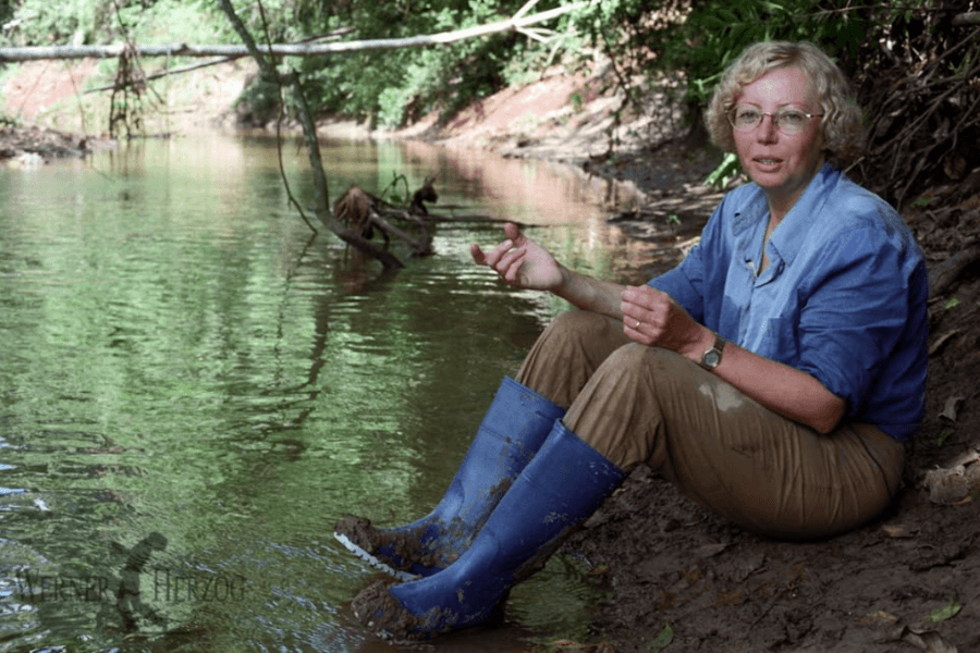 A woman in a blue shirt, muddy khaki pants, and bright blue boots sits by a riverbank in a tropical forest. She gestures with her hands while speaking, with dense greenery and calm water surrounding her.