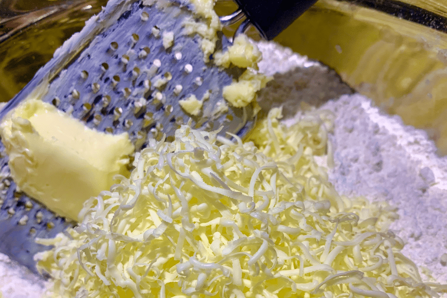Close-up of cold butter being grated into a bowl of flour using a box grater, creating fine shreds of butter for a baking recipe. This technique helps evenly distribute the butter for flaky doughs like biscuits or pie crust.