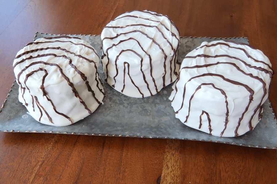 Three large, homemade desserts styled to look like classic Hostess Zingers are placed on a metal tray. Each is coated in a thick layer of white icing and decorated with curved chocolate drizzle lines mimicking the signature swirl pattern.