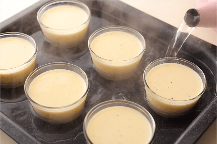 Several small glass ramekins filled with a custard mixture are arranged in a baking tray while hot water is being poured around them to create a water bath (bain-marie), a gentle baking method used for even cooking.