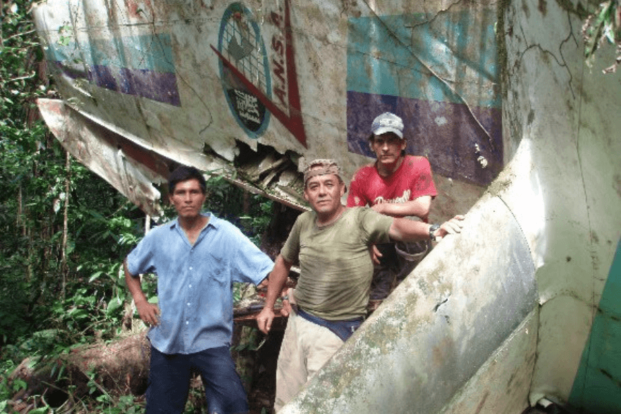 Three men pose in front of a moss-covered, rusted airplane wreck deep in the jungle, with part of the fuselage showing the logo and faded colors of the airline LANSA. The dense forest surrounds the damaged tail section, which bears visible signs of age and decay.