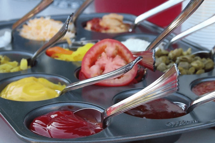Muffin tin repurposed as a condiment and topping tray, holding various ingredients such as ketchup, mustard, barbecue sauce, sliced tomatoes, cheese, pickles, onions, and more. Each compartment has a spoon for serving.