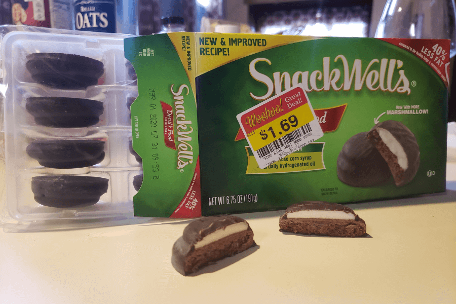 A box of SnackWell’s Devil’s Food cookies sits next to a plastic tray containing the chocolate-coated marshmallow treats. Two cookies are cut in half in front, showing the soft chocolate cake and fluffy white marshmallow filling. The box is labeled with a sale sticker for $1.69 and boasts a “new & improved recipe.”