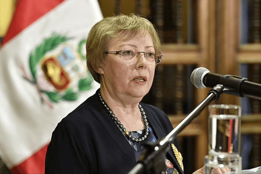 A woman wearing glasses and a dark blazer speaks into a microphone at a podium, with a glass of water in front of her. Behind her is the national flag of Peru, suggesting an official or diplomatic event.
