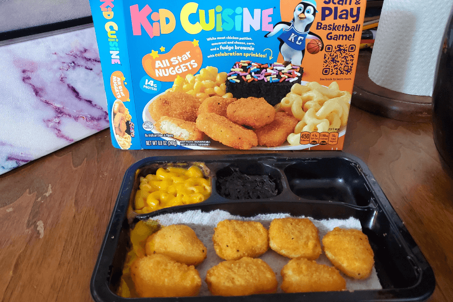 A Kid Cuisine frozen meal with five breaded chicken nuggets, a portion of creamy macaroni and cheese, and a chocolate fudge brownie in a black plastic tray. The colorful box in the background features the “All Star Nuggets” label, corn, mac and cheese, and a brownie with rainbow sprinkles, alongside a cartoon penguin mascot.