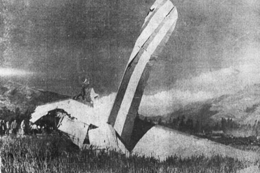 Black-and-white photo showing the wreckage of an airplane crash, with the vertical stabilizer featuring red and white stripes sticking out from a destroyed fuselage in a grassy field. People can be seen in the background examining the crash site.