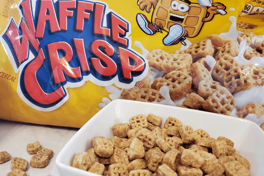 A close-up of a bowl filled with Waffle Crisp cereal placed in front of a large, colorful cereal bag. The packaging features the brand name in bold red and blue letters, along with a cartoon waffle character and a splash of milk surrounding the mini waffle-shaped cereal pieces.