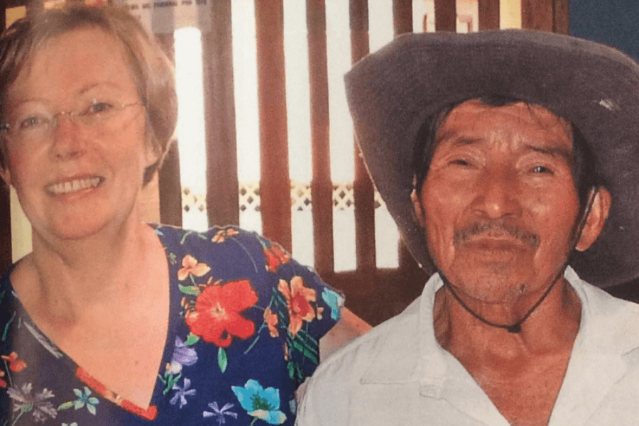 A smiling woman in a colorful floral shirt stands beside an older man wearing a wide-brimmed hat and a white shirt. They appear friendly and relaxed, posing indoors with wooden paneling in the background.