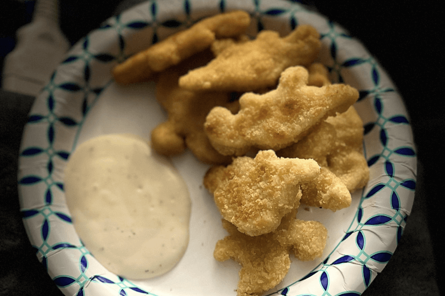 A paper plate holding several dinosaur-shaped chicken nuggets with a side of ranch dressing. The nuggets are golden brown and crispy, shaped like different dinosaurs including a T-Rex and a Stegosaurus.