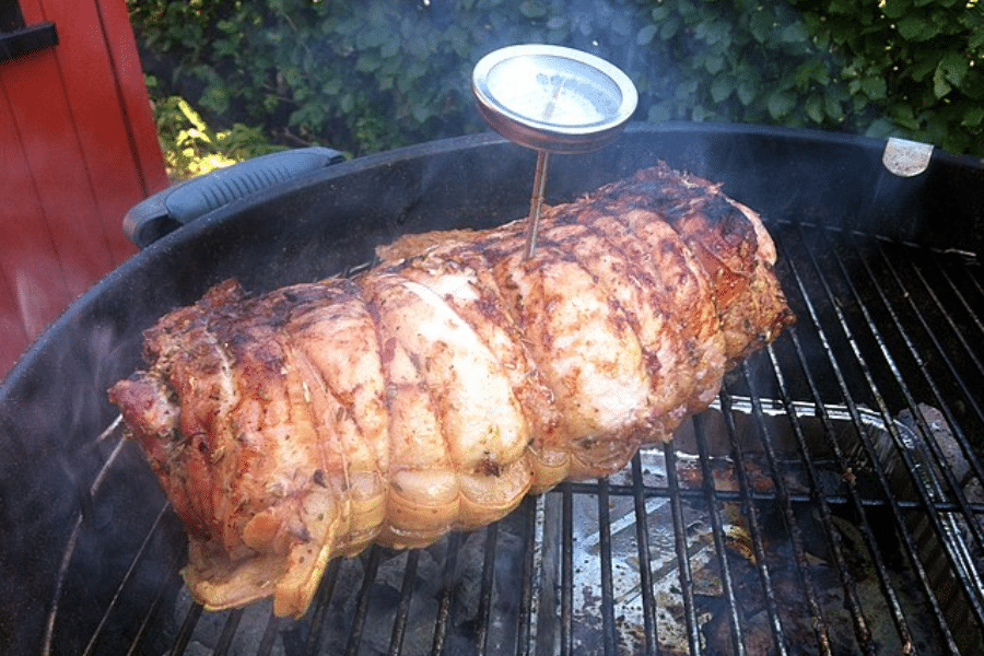 Rolled and tied roast cooking on a charcoal grill with smoke rising and a meat thermometer inserted into the center to monitor internal temperature. The roast is golden brown and crisp on the outside, indicating slow, even cooking.