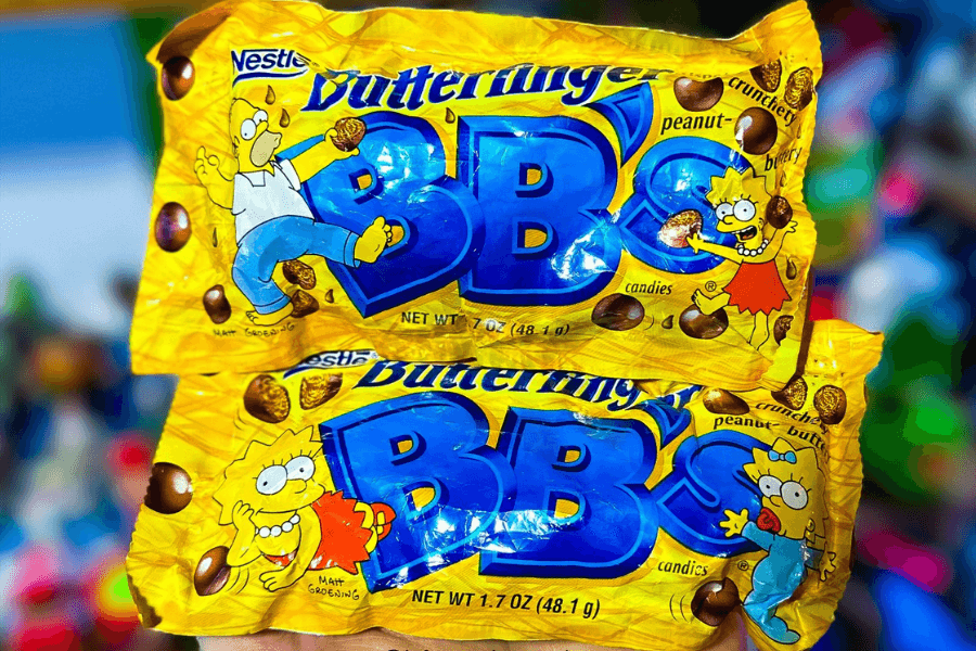Two nostalgic yellow bags of Nestlé Butterfinger BB’s candies, featuring characters from The Simpsons. The top bag showcases Homer and Lisa Simpson holding the round, crunchy peanut-buttery candies, while the bottom bag features Lisa and baby Maggie Simpson next to the BB’s logo.