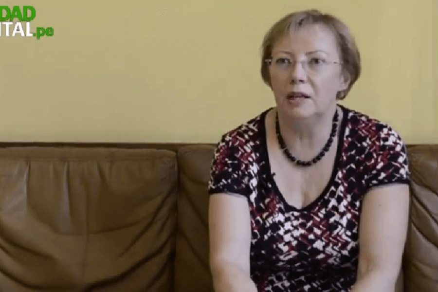 A woman with short hair and glasses sits on a brown leather couch against a pale yellow wall, speaking mid-sentence during what appears to be an interview. She wears a patterned red, black, and white top and a beaded necklace, with a calm yet focused expression.