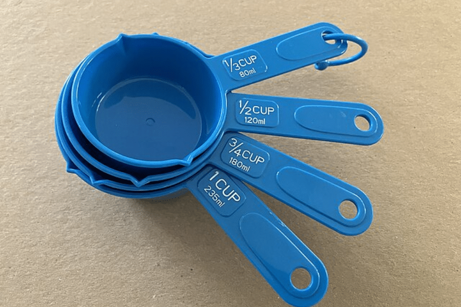 Set of four blue plastic measuring cups labeled with both cup and milliliter measurements, fanned out on a beige surface. The sizes include 1 cup (235ml), 3/4 cup (180ml), 1/2 cup (120ml), and 1/3 cup (80ml), all connected by a plastic ring.