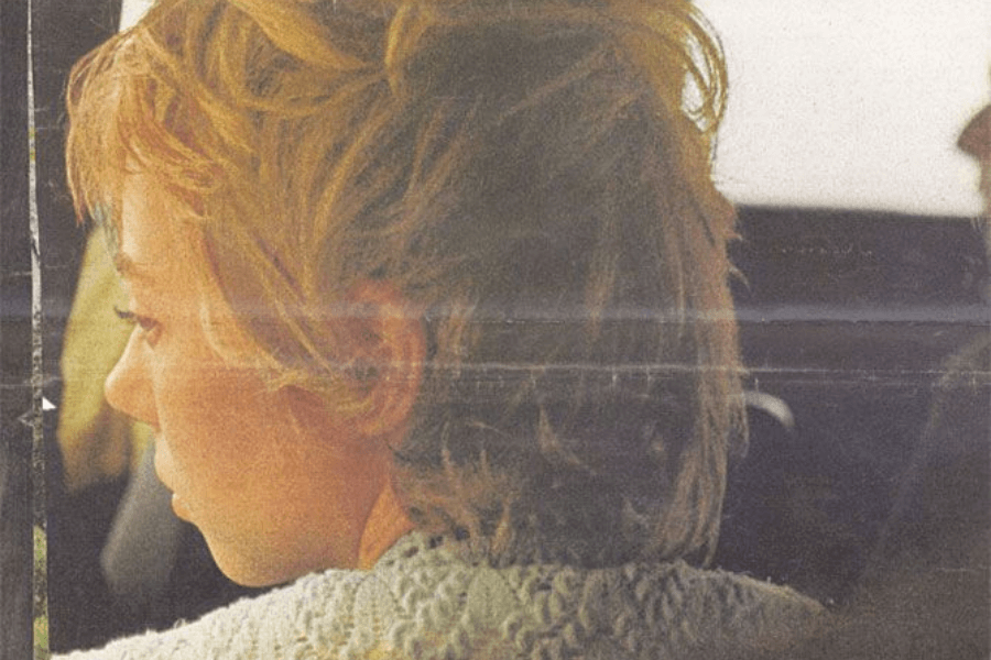 A woman with tousled blonde hair, wearing a textured sweater, is seen in profile looking down or out a window from inside a vehicle. The image has a vintage, grainy quality, adding a nostalgic or reflective mood.