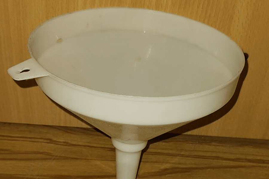 White plastic funnel with a wide opening and narrow spout, placed upright on a wooden surface against a wood-paneled background. A small tab with a hole on the rim allows for hanging storage.