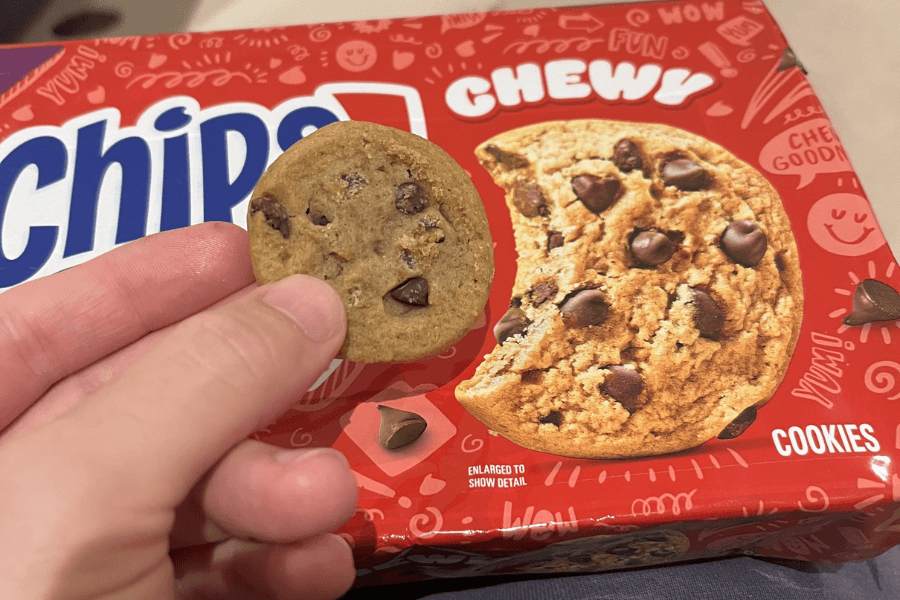 Close-up of a hand holding a small chewy Chips Ahoy chocolate chip cookie in front of a red box of Chips Ahoy Chewy Cookies. The packaging features a large image of a soft, gooey chocolate chip cookie with fun doodles and words like “yum” and “chewy” scattered across the background.