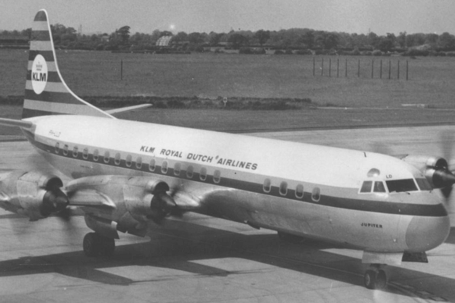 Black and white photo of a vintage KLM Royal Dutch Airlines propeller aircraft on a runway, labeled "JUPITER" near the nose. The plane features four engines and a streamlined design, with open grass fields in the background.