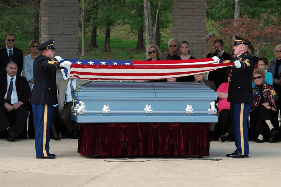 Two uniformed military personnel solemnly fold an American flag over a closed casket during a funeral service, surrounded by seated mourners. The scene takes place outdoors under a pavilion, with a forested backdrop, paying tribute to a fallen veteran with full military honors.