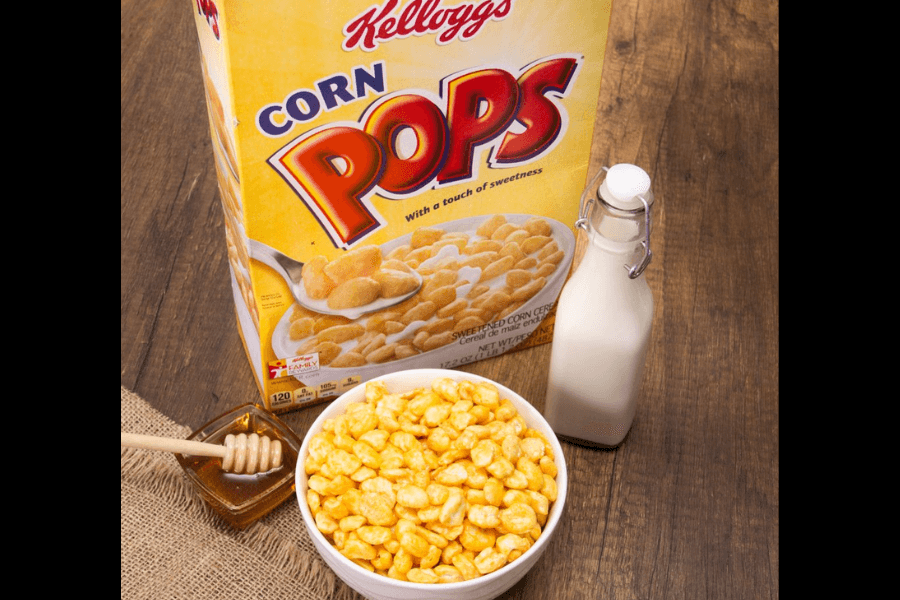 Box of Kellogg's Corn Pops cereal next to a white bowl filled with the golden puffed corn cereal, a glass bottle of milk, and a small dish of honey with a honey dipper on a wooden table.
