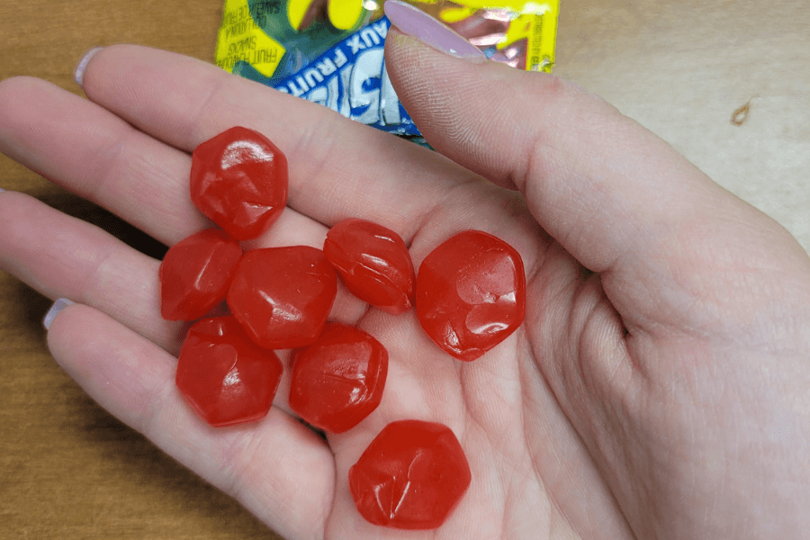 A hand holding a handful of red gummy candies, shaped like raspberries or jewels, with part of a colorful "Juicy Burst" fruit snack package visible in the background.