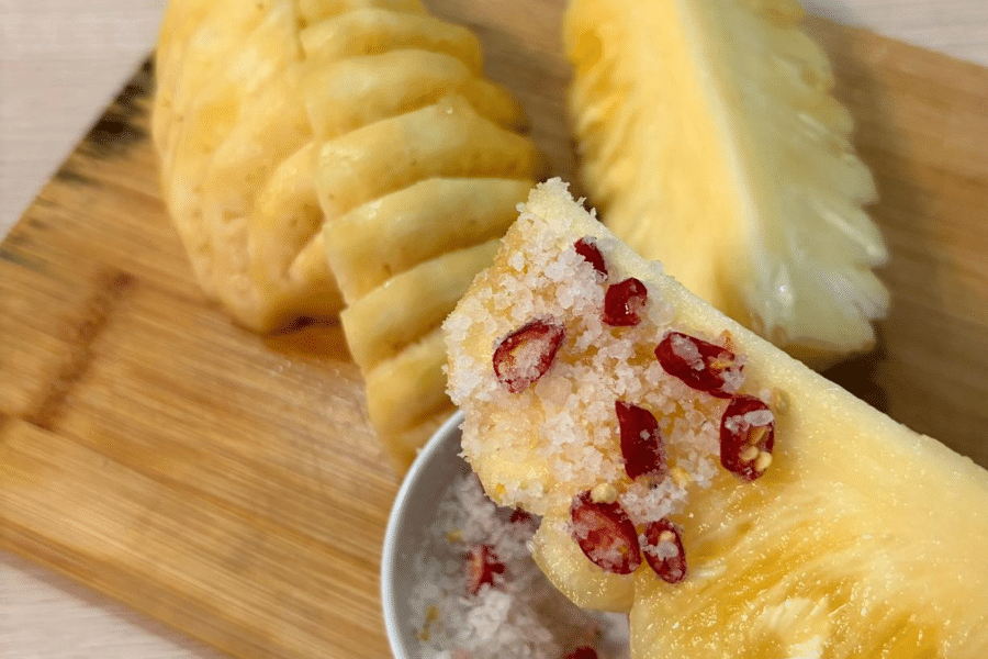 Close-up of a fresh pineapple wedge dipped in a mixture of coarse salt and sliced red chili peppers, placed over a small bowl of the same mixture. In the background, other peeled and sliced pineapple pieces rest on a wooden cutting board.