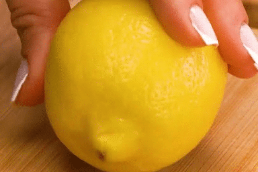 Close-up of a bright yellow lemon being gently rolled on a wooden surface by a hand with neatly manicured white nails. This common technique helps release more juice from the lemon before cutting.