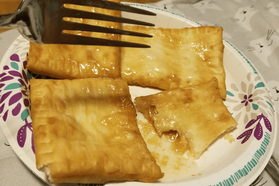A paper plate with three golden-brown, glazed rectangular pastries and a fourth piece partially bitten to reveal a gooey, syrupy filling inside. A metal fork is held above the plate, ready to pick one up.