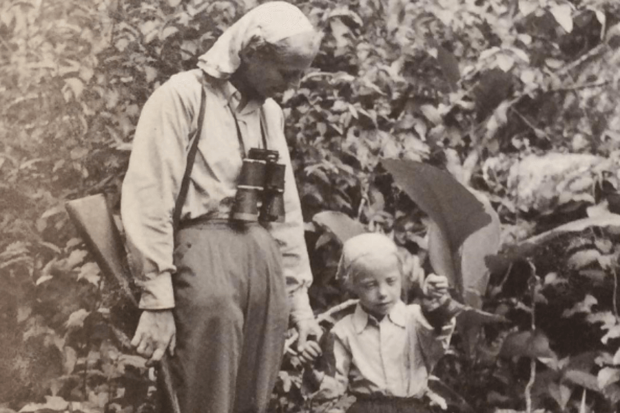 Black-and-white photo of an older woman and a young blonde child in a dense jungle. The woman wears a headscarf, has binoculars around her neck, and carries a rifle over her shoulder. The child looks curiously at a large leaf they are holding.