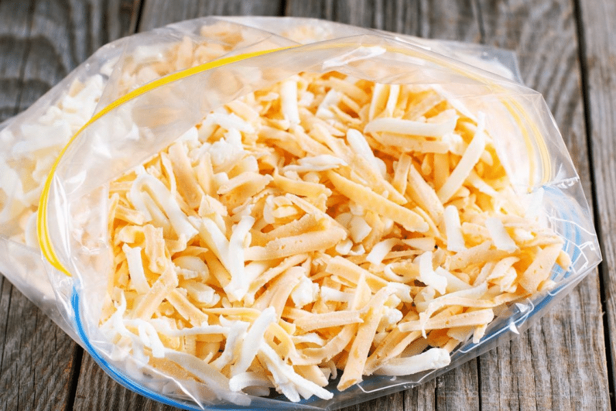 Clear zip-top freezer bag filled with a mix of shredded white and orange cheese, resting on a rustic wooden surface. The cheese appears freshly grated and ready for storage or meal prep.