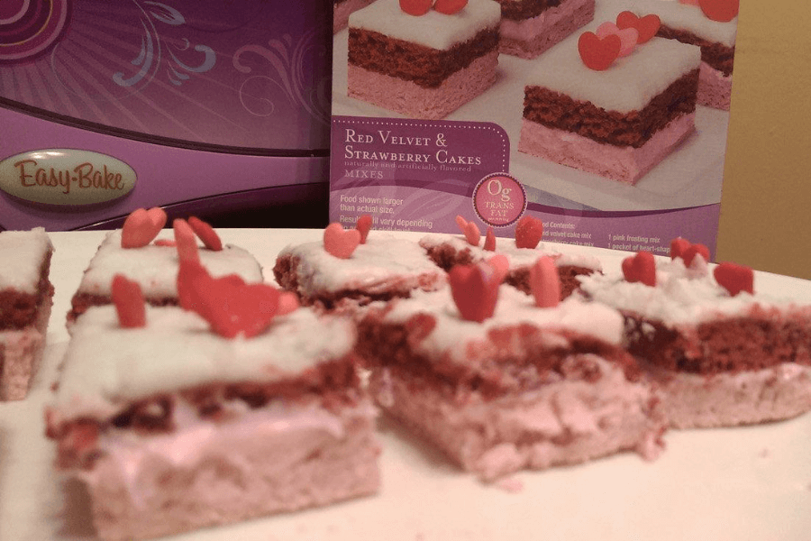 Miniature red velvet and strawberry cake squares topped with white frosting and heart-shaped red and pink sprinkles, displayed on a plate in front of the Easy-Bake oven and the original cake mix box labeled "Red Velvet & Strawberry Cakes."
