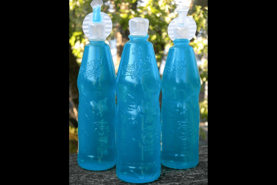 Three translucent blue Kool-Aid Bursts bottles with twist-off caps, lined up outdoors on a wooden surface. The bottles are filled with a bright blue drink and feature embossed fruit graphics and the Kool-Aid logo.