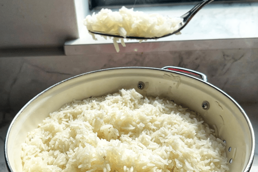Freshly cooked white rice in a large stainless steel pot, with a spoonful of rice being held above the pot near a sunlit window. The rice appears fluffy and well-cooked, with individual grains separated.