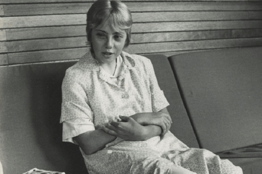 Black-and-white photo of a young woman sitting on a cushioned bench with wooden paneling in the background. She wears a light-patterned dress and has her arms crossed over her midsection, appearing to be in conversation or deep thought.